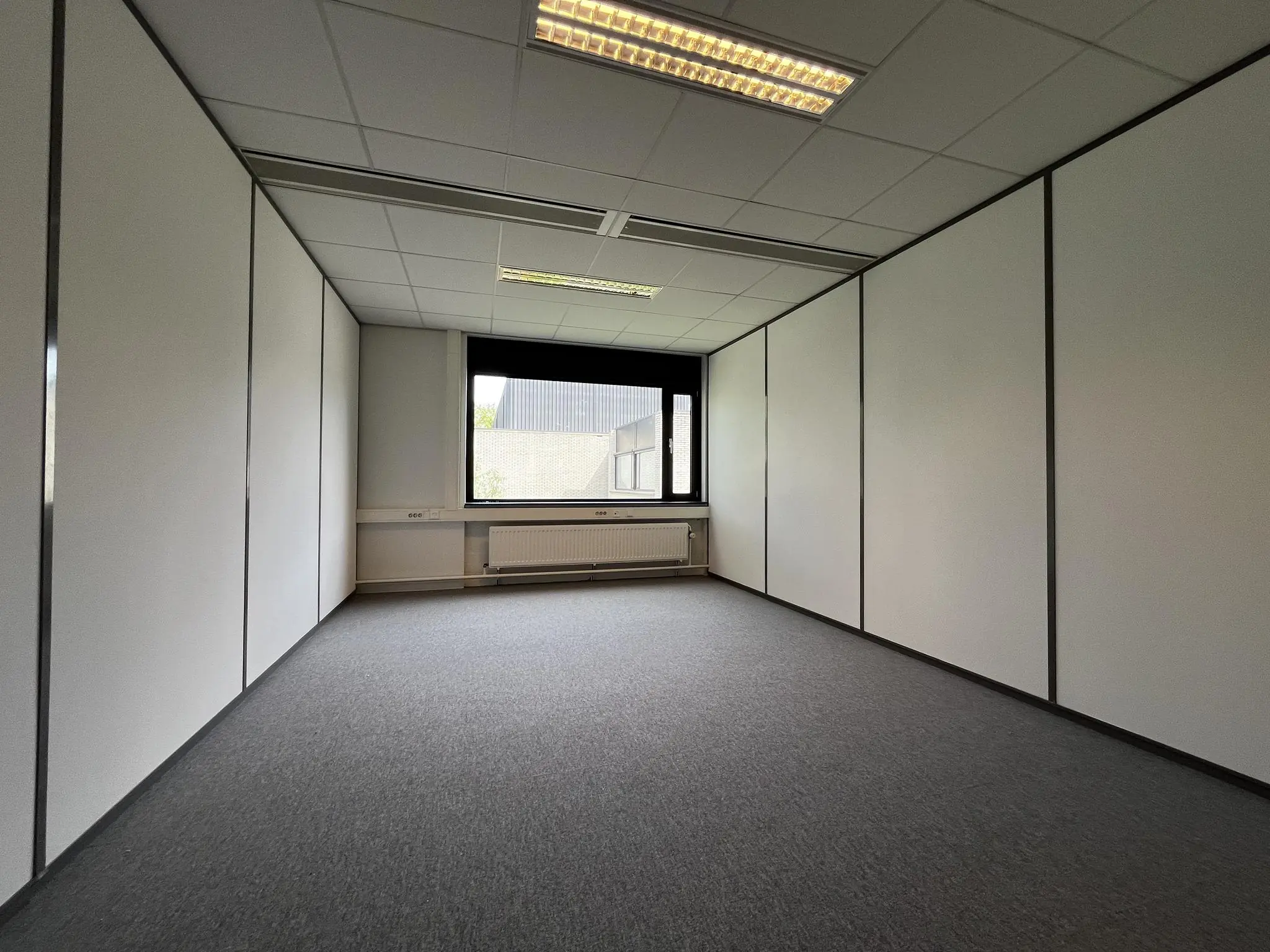 Leeg kantoor met systeemplafond, tapijtvloer en groot raam aan de achterkant in gebouw aan de Burgemeester Verderlaan.